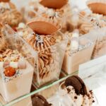 004ba70ef25f7cdf743eba971a333df2jpg › Udanewypieki Assorted cream-based desserts in clear square containers with decorative swirls and chocolate pieces on top.