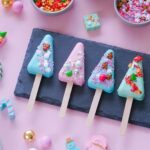 00b893c3b9067a44fc267647e529942bjpg › Udanewypieki Four pastel-colored, tree-shaped popsicles on a slate serving board, decorated with sprinkles and candy decorations against a pink background.