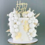 White cake surrounded by white balloons and gold leaf accents, with a cream 'Happy Birthday' topper on top of the cake boundary edge.