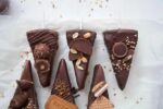 3b700d7692ffaaadfd531663f1efb638jpg › Udanewypieki Row of chocolate-dipped ice cream bars on sticks with toppings like peanuts, crushed cookies, and wafers on parchment tailing crumbs nearby.