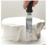 3c2ad393336c3f1fe484affe80cac0c9jpg › Udanewypieki Close-up of a hand guiding a knife to cut a white frosted cake on a plate.