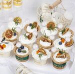 Assorted white-frosted cupcakes decorated with dried orange slices, blueberries, rosemary, and decorative picks on a white display table for a party favors setup