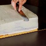 6d5d3e8533c1ac4c3c6092ab9d9906b2jpg › Udanewypieki Person smooths gray frosting over a large rectangular cake with an offset spatula in hand.