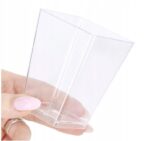 981ffc0a8f96c620c28a5abd85d8fa47jpg › Udanewypieki Hand holding a clear disposable plastic cup between thumb and forefinger, with light pink nails visible on the thumb side.