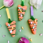 f9350220b7046932f83703e84bbd293bjpg › Udanewypieki Orange carrot-shaped cookies on sticks with green icing and candy sprinkles on a pastel green background